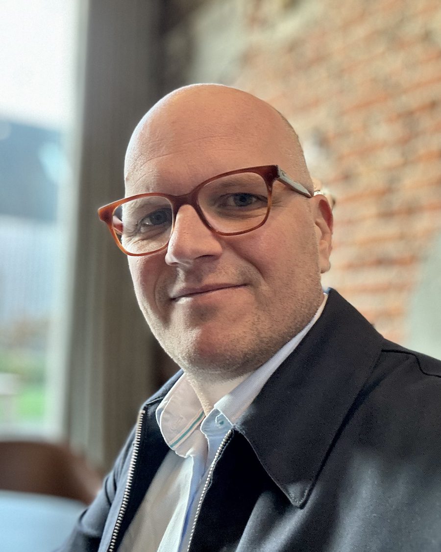 Jan Jacobs, glimlachend aan het raam, met een bakstenen muur achter hem